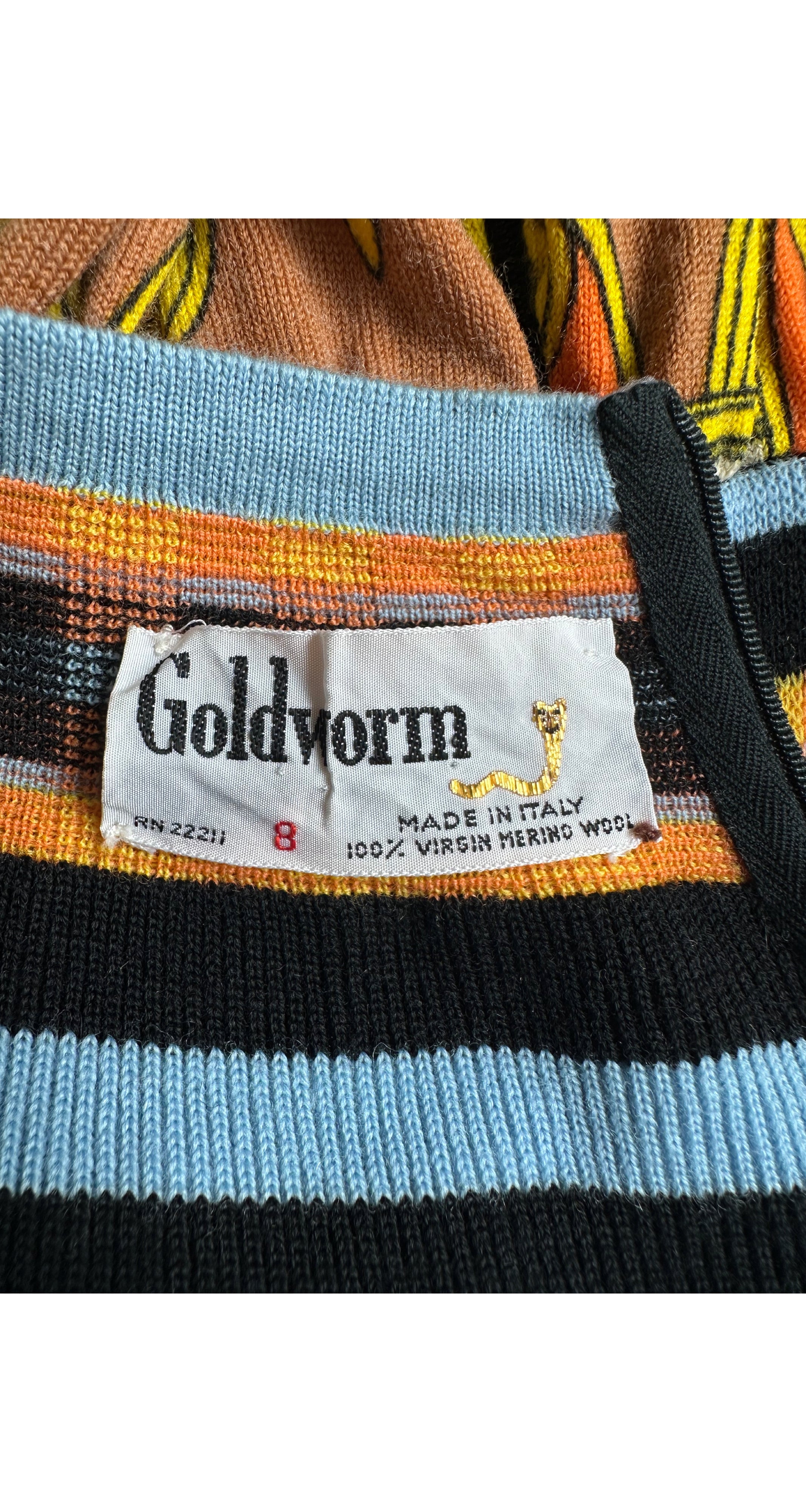 Close-up of a Goldworm knit garment showing a striped pattern with black, light blue, orange, and brown horizontal bands, along with a white brand label.