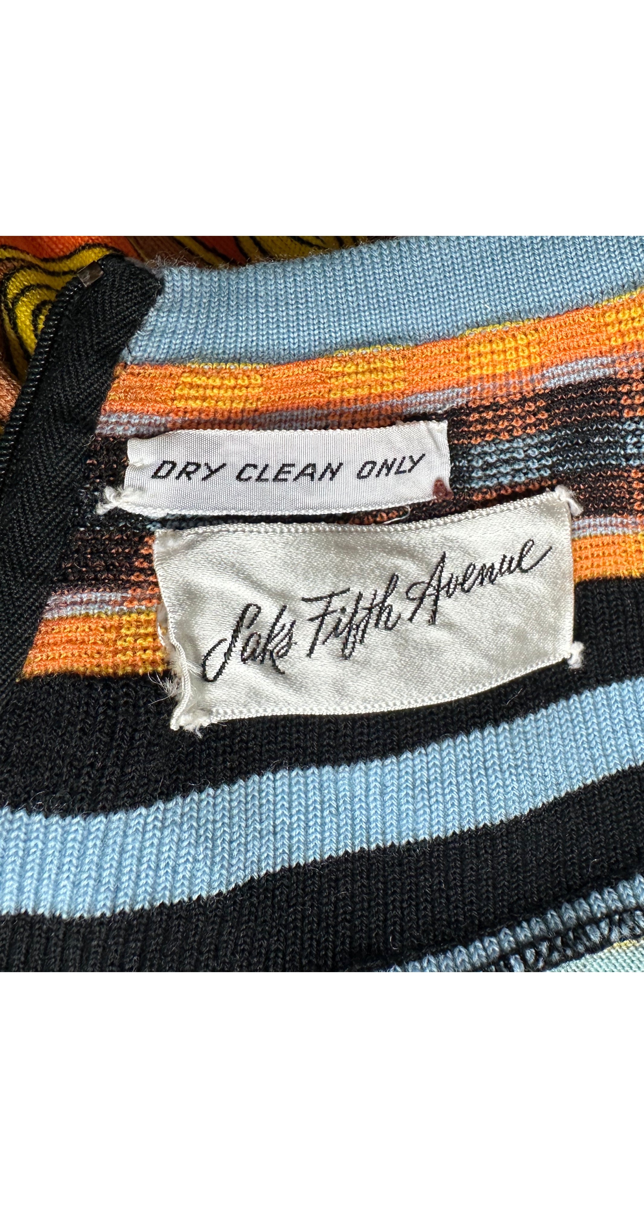 Close-up of a striped knit garment with black, light blue, orange, and brown stripes, showing two labels: one reading 'DRY CLEAN ONLY' and the other 'Saks Fifth Avenue'.