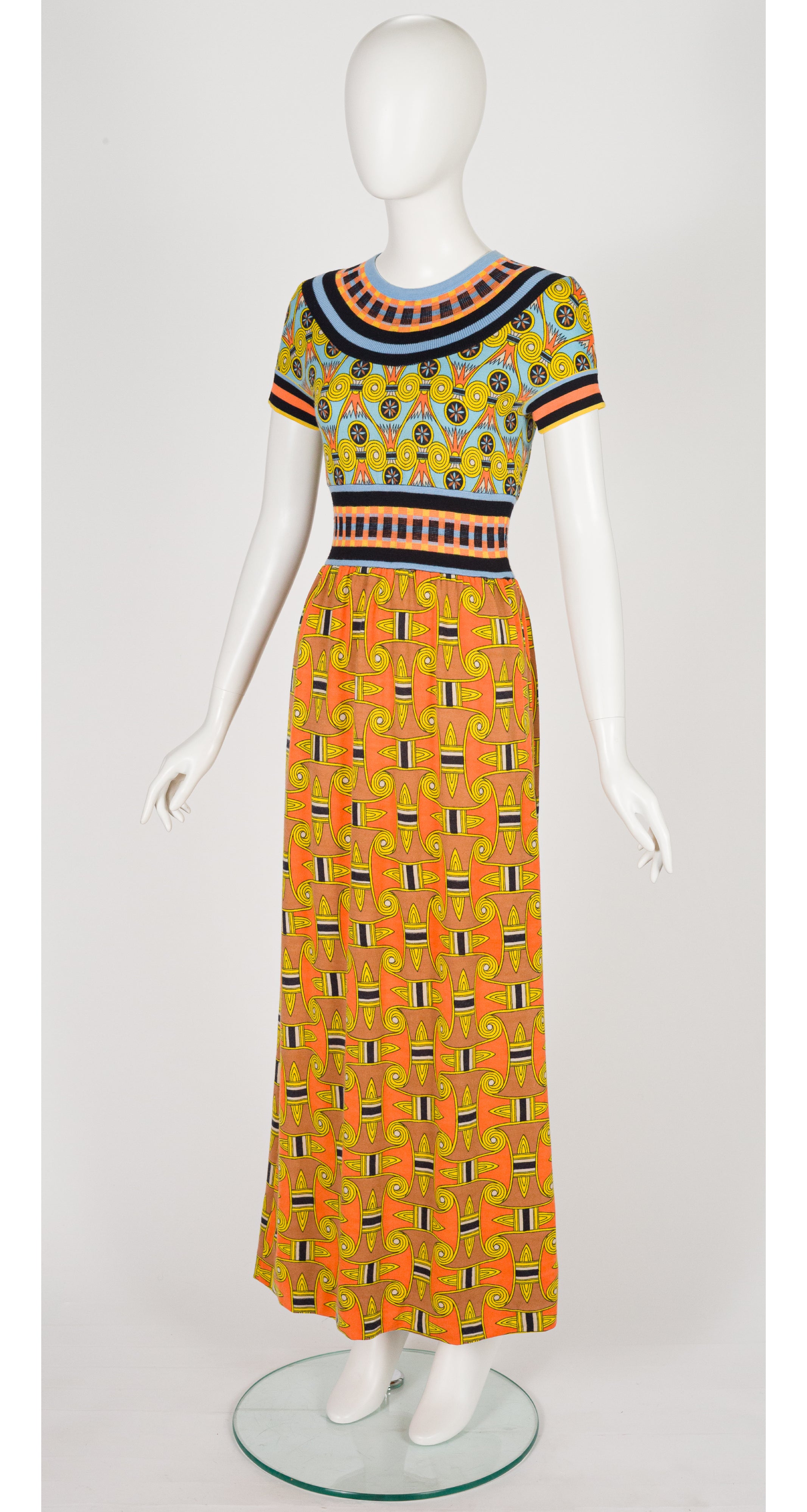 A 1970s Goldworm short-sleeve maxi dress displayed on a white mannequin, featuring a vibrant orange and yellow patterned design with black, blue, and green accents.