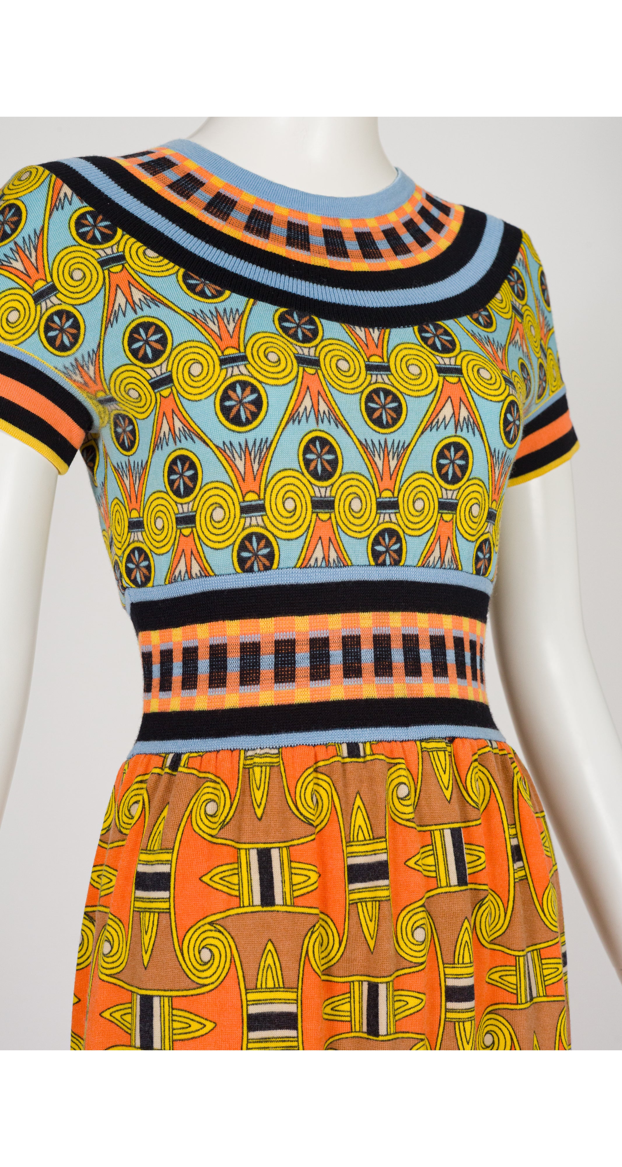 A 1970s Goldworm short-sleeve dress displayed on a white mannequin, featuring a vibrant split design with a light blue and yellow geometric pattern on the upper bodice and a bright orange and yellow patterned skirt.