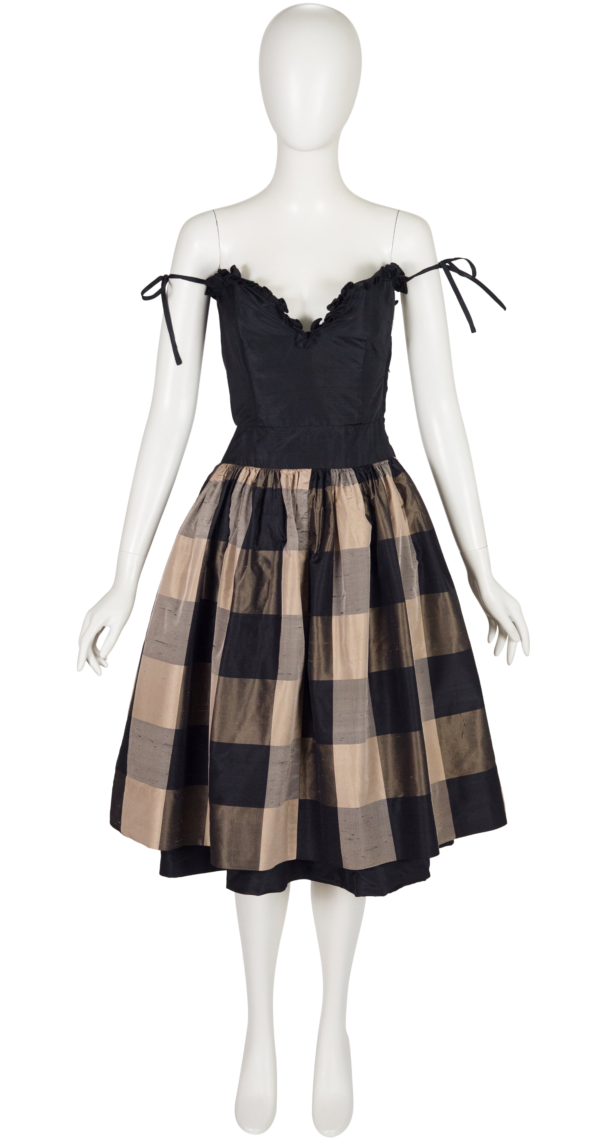 A black and beige Valentino 1982 SS plaid dress displayed on a white mannequin. The dress features a black corset-style bodice with ruffles and tie straps, and a full pleated skirt in a black and beige check pattern.