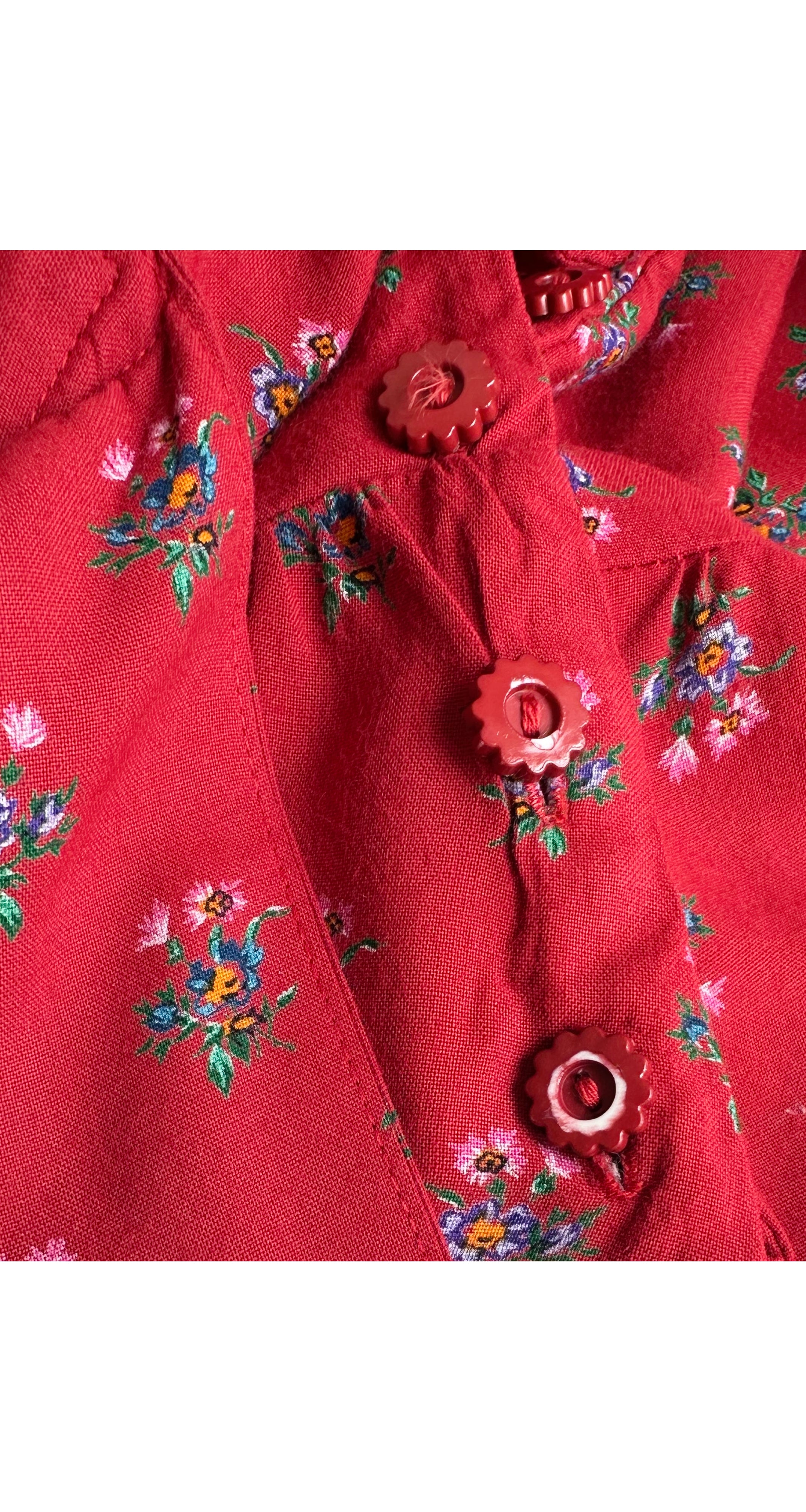 A close-up of a red fabric garment featuring a small-scale floral print in green, blue, and pink, detailed with matching red decorative buttons.