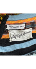 Close-up of a striped knit garment with black, light blue, orange, and brown stripes, showing two labels: one reading 'DRY CLEAN ONLY' and the other 'Saks Fifth Avenue'.