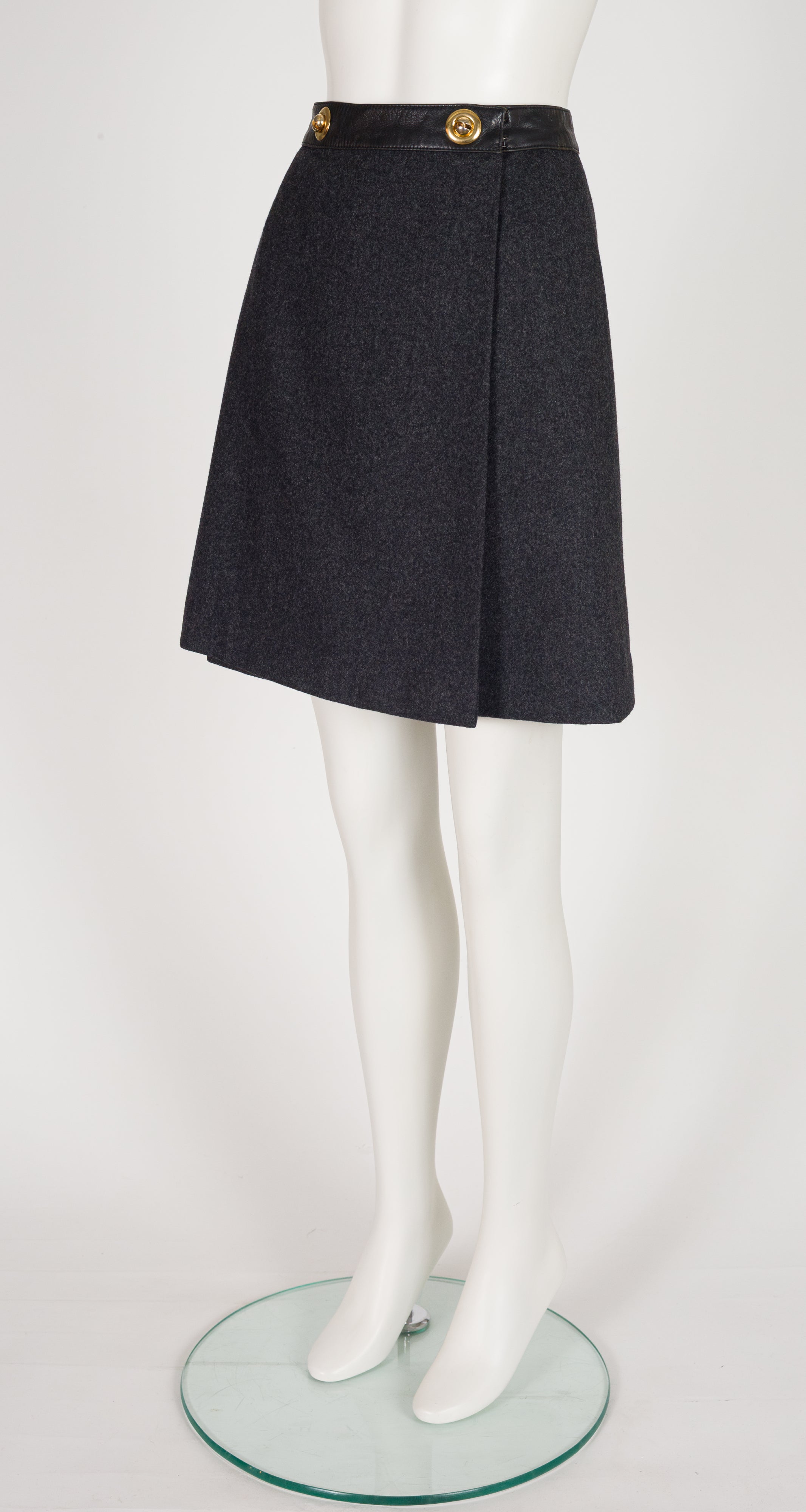 A 1970s Hermes dark gray textured A-line skirt with a black waistband and gold-tone buttons, displayed on a white mannequin leg form over a round glass base.