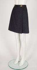A 1970s Hermes dark gray textured A-line skirt with a black waistband and gold-tone buttons, displayed on a white mannequin leg form over a round glass base.