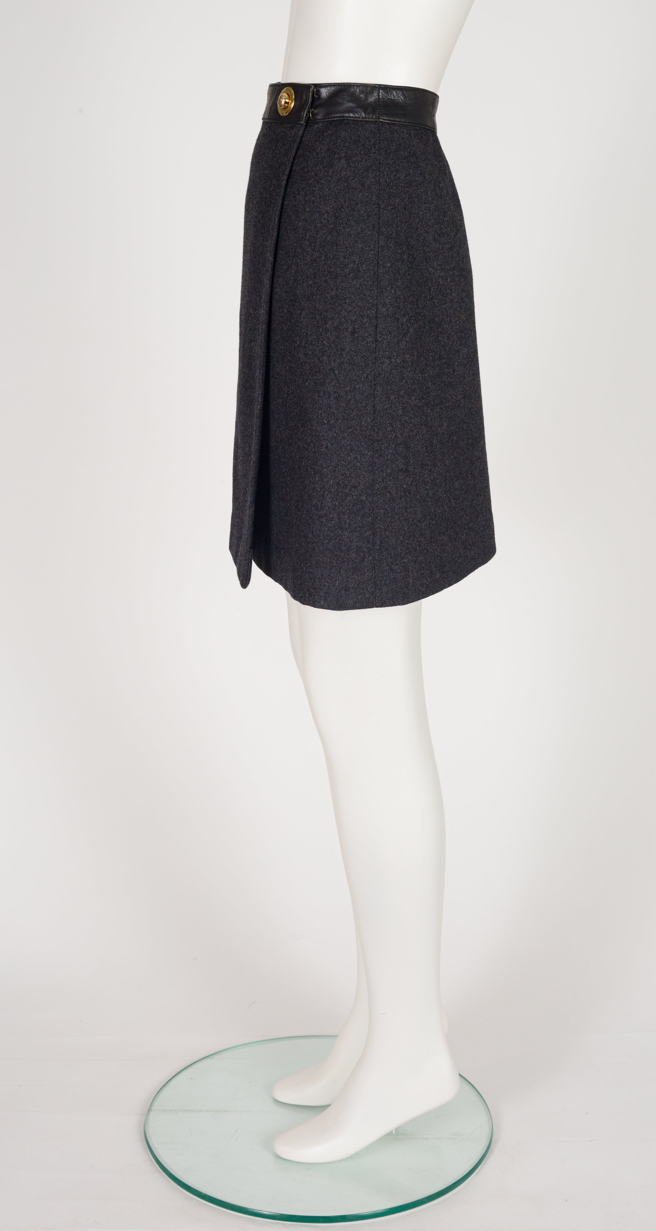 A 1970s Hermes dark charcoal gray wrap-style skirt with a black leather waistband and a gold button, displayed on a white mannequin leg stand.