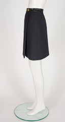 A 1970s Hermes dark charcoal gray wrap-style skirt with a black leather waistband and a gold button, displayed on a white mannequin leg stand.