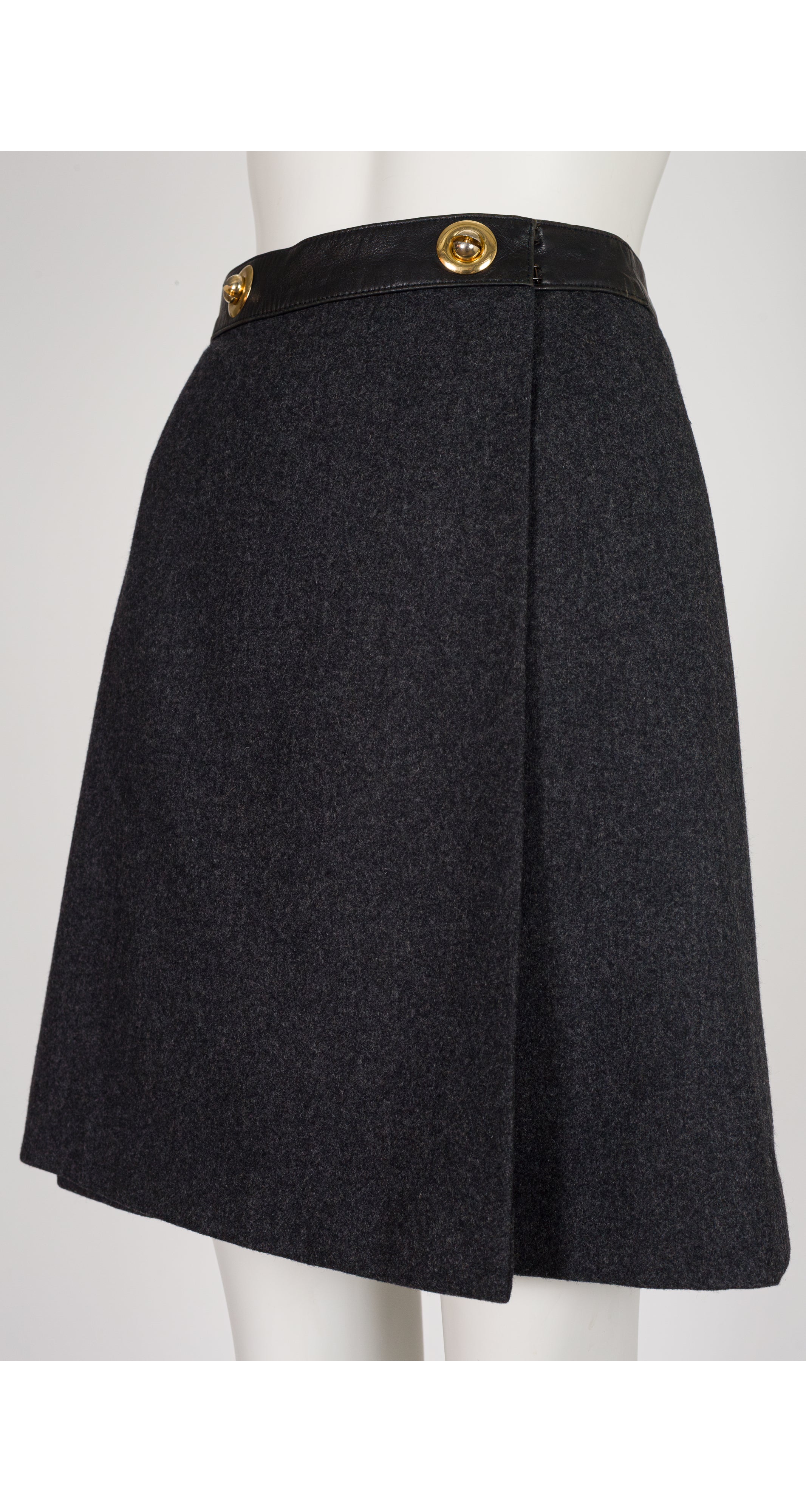 A 1970s Hermes dark charcoal gray wool skirt with a black waistband and two prominent gold-tone buttons, displayed on a white mannequin torso.
