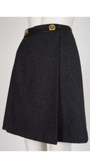 A 1970s Hermes dark charcoal gray wool skirt with a black waistband and two prominent gold-tone buttons, displayed on a white mannequin torso.