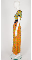 A 1970s Goldworm short-sleeve maxi dress displayed on a white mannequin, featuring a vibrant yellow, orange, green, and blue geometric pattern with a striped waistband.