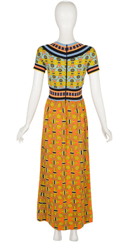 A 1970s Goldworm short-sleeve maxi dress displayed on a white mannequin, featuring a vibrant orange and yellow patterned skirt with a matching patterned bodice and striped trim.