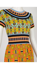 A 1970s Goldworm short-sleeve dress displayed on a white mannequin, featuring a vibrant split design with a light blue and yellow geometric pattern on the upper bodice and a bright orange and yellow patterned skirt.