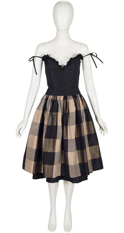 A black and beige Valentino 1982 SS plaid dress displayed on a white mannequin. The dress features a black corset-style bodice with ruffles and tie straps, and a full pleated skirt in a black and beige check pattern.