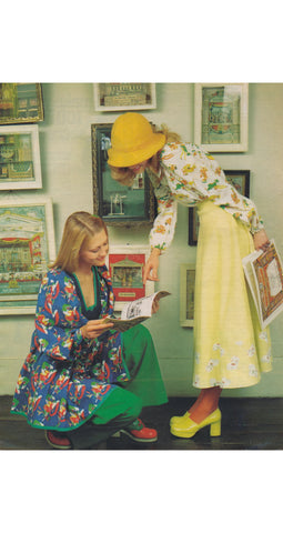 Two women in Jeff Banks colorful retro-style clothing pose in front of a wall covered in framed pictures. One woman is kneeling and wearing a blue floral robe, while the other stands wearing a yellow hat, a white floral blouse, a pale yellow skirt, and yellow high heels.